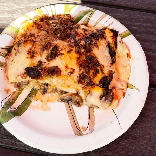 Cheesy, browned lasagna with mushrooms on a disposable plate, served with a small cup of creamy sauce on a dark wooden table.