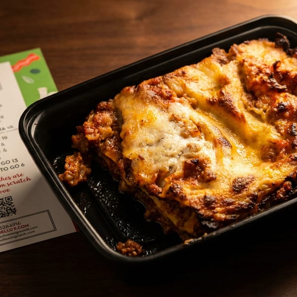 Cheesy lasagna in a black takeout tray beside a Mattiello’s menu on a wooden table.