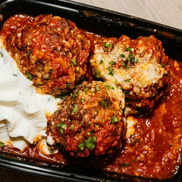 Three meatballs topped with tomato sauce, melted cheese, and herbs, served with sour cream in a black dish.