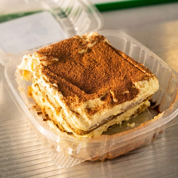 Square slice of tiramisu dusted with cocoa in an open plastic takeaway container on a wooden table.