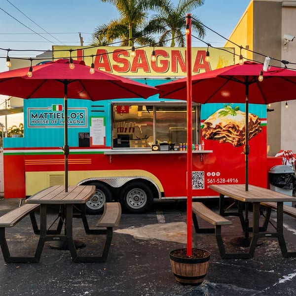 Colorful Mattiello’s House of Lasagna food trailer with red umbrellas, string lights, and picnic tables outside.